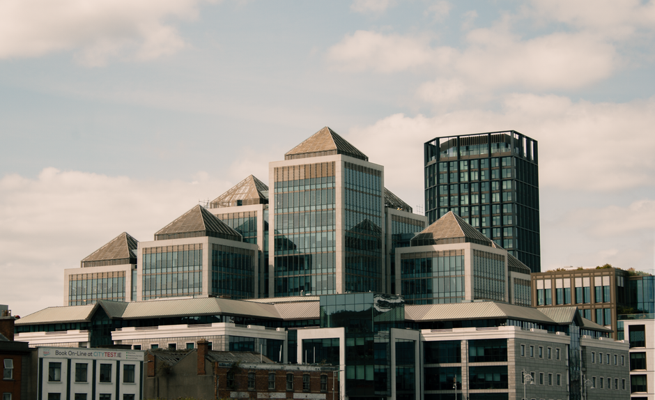 A modern office building with multiple glass facades and geometric roof structures is situated in an urban setting under a partly cloudy sky. The building features several interconnected sections with