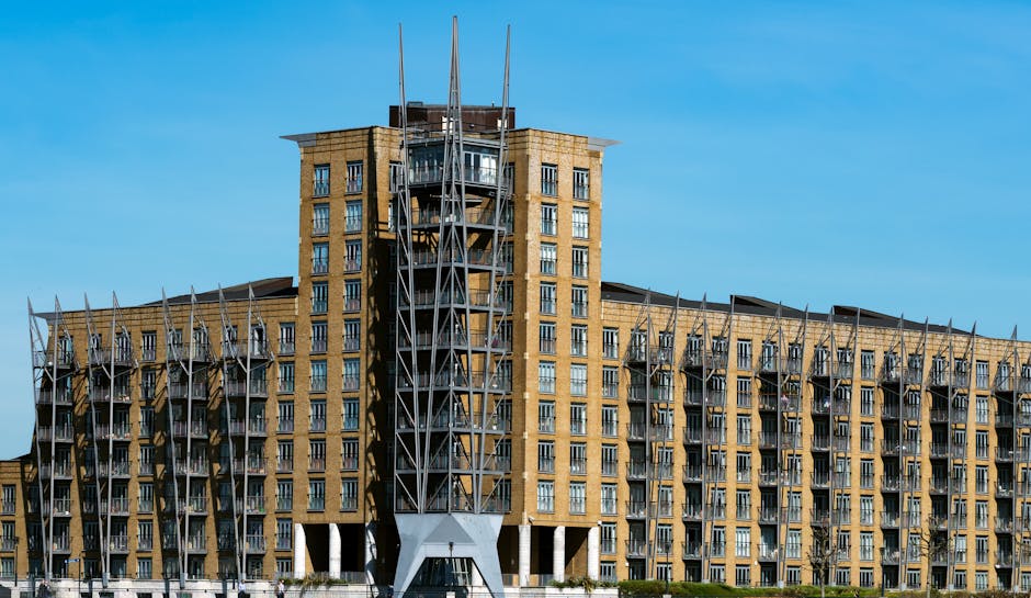 A modern office building with multiple glass facades and geometric roof structures is situated in an urban setting under a partly cloudy sky. The building features several interconnected sections with